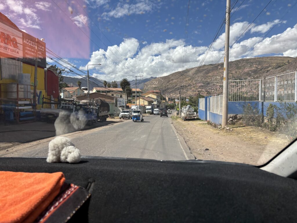 A view from inside a vehicle looking out onto a road in a rural area. The road is lined with buildings on the left, including a colorful structure with signs. On the right, there is a blue fence and some vegetation. In the distance, there are hills under a partly cloudy sky. The dashboard of the vehicle has a small white fluffy object and an orange cloth. Several vehicles are visible on the road ahead.