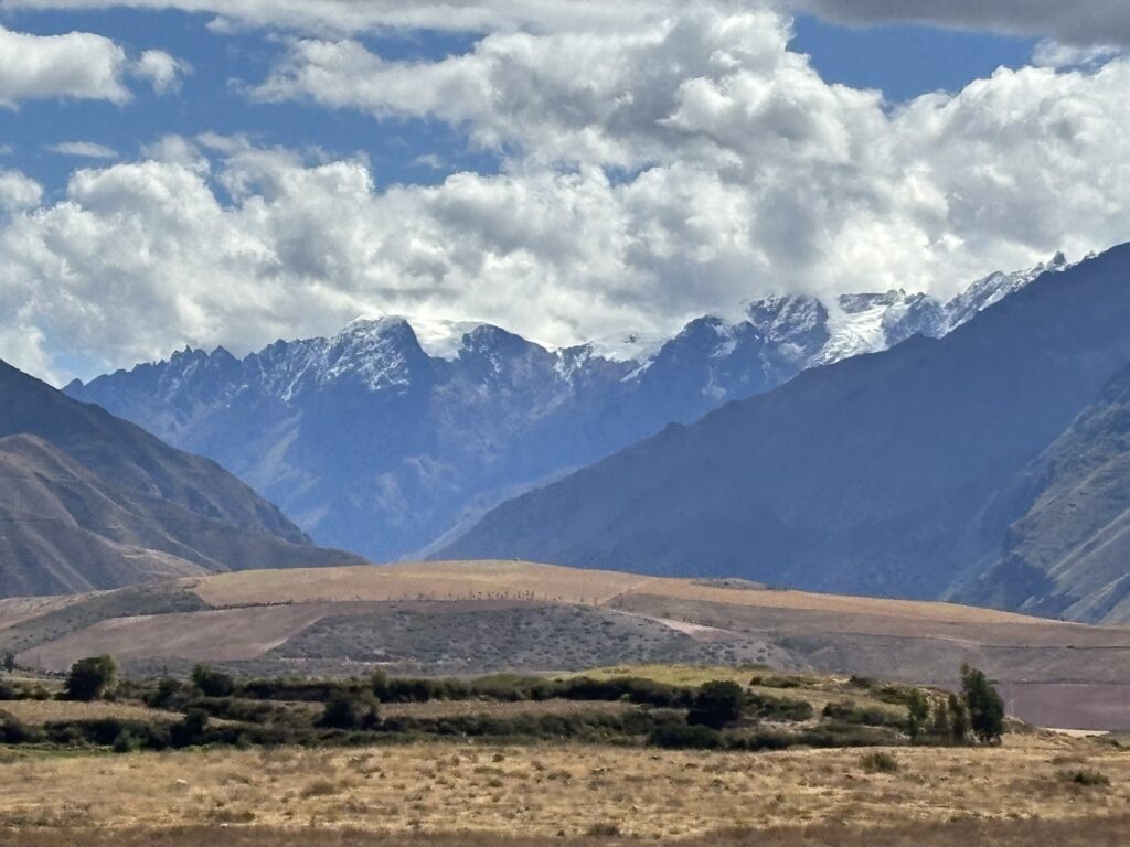 A scenic landscape featuring a range of mountains with snow-capped peaks under a partly cloudy sky. In the foreground, there are fields and patches of greenery, with rolling hills leading up to the mountains. The overall atmosphere is serene and natural.