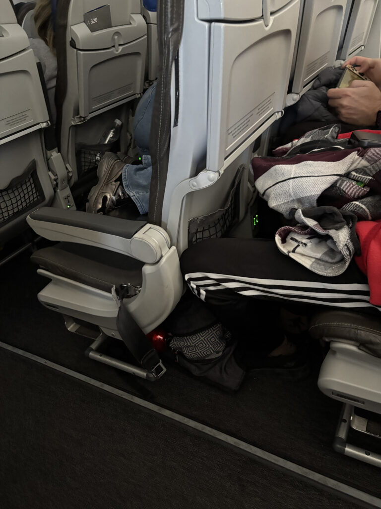 The image shows the interior of an airplane cabin, focusing on the seating area. A person is seated on the right, wearing black pants with white stripes, holding a phone. There are various items, including a jacket and a scarf, on their lap. The seat in front has a person with their legs crossed, wearing jeans and sneakers. The floor area has a bag and a red object, possibly a bottle. The seats have tray tables and seatback pockets.
