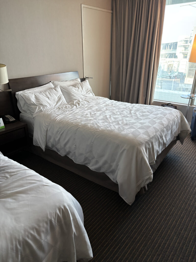 A hotel room with two neatly made beds covered in white linens. The room has a window with closed curtains on the right side, allowing some natural light to enter. A bedside table with a lamp and a telephone is visible next to one of the beds. The floor is carpeted with a striped pattern.