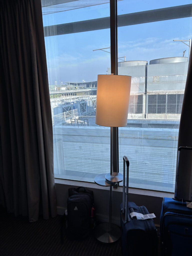 A hotel room with a large window showing an industrial rooftop view. Inside, there is a floor lamp with a white shade next to a small round table. Two pieces of luggage, a backpack and a suitcase, are placed near the window. The curtains are partially drawn to the side.