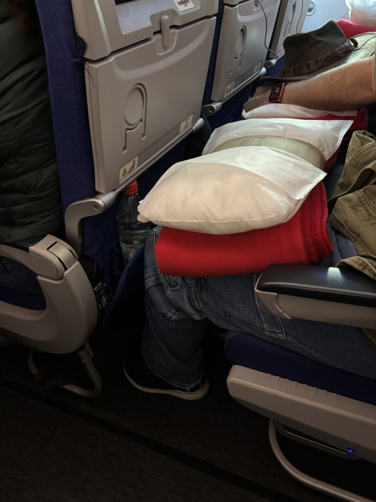 A person is seated on an airplane, wearing jeans and a watch. They have a red blanket and a white pillow on their lap. A hat is placed on the armrest. The seatback tray tables are closed, and a water bottle is visible in the seat pocket.