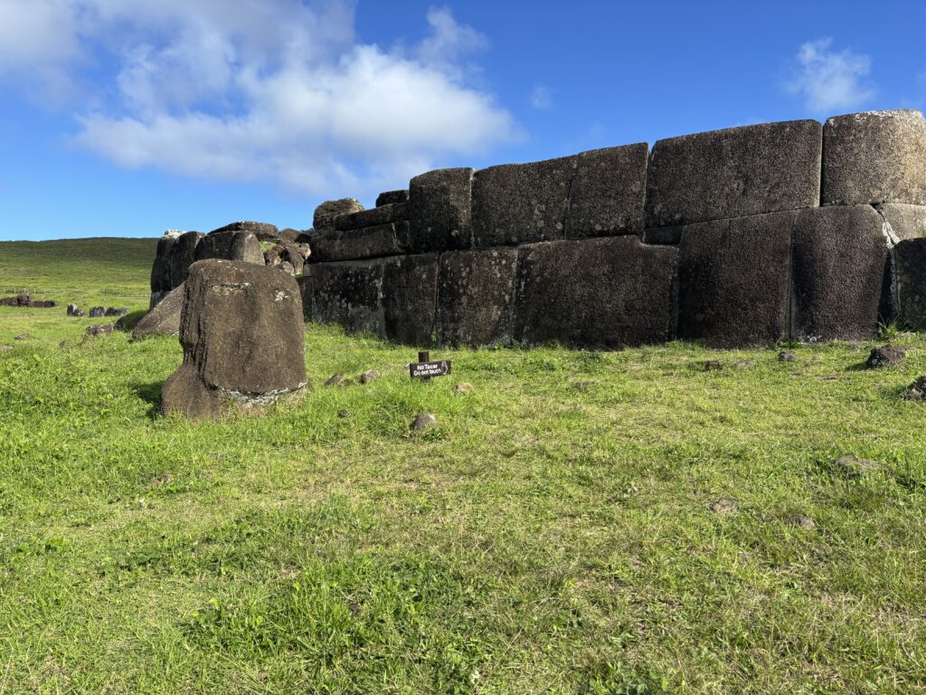 The image shows a section of an ancient stone wall made of large, rectangular stones stacked together. The wall is situated on a grassy field under a blue sky with some clouds. A small sign is visible in front of the wall.