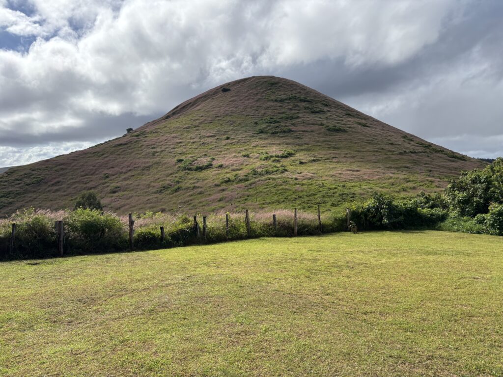 A grassy hill under a cloudy sky. The hill is covered with patches of green and brown vegetation. In the foreground, there is a grassy field bordered by a simple wooden fence with some bushes. The sky is partly cloudy, with sunlight filtering through.
