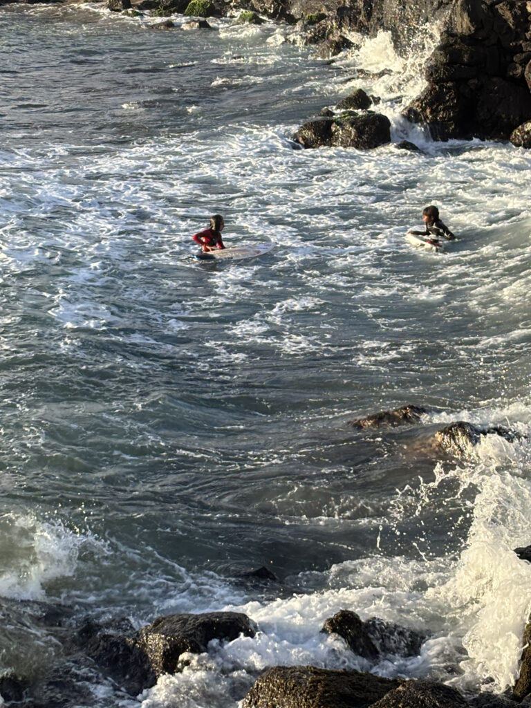 Two people are in the ocean near rocky cliffs, each on a surfboard. The water is choppy with waves crashing against the rocks. One person is wearing a red wetsuit, and the other is in a black and white wetsuit. The scene captures an active moment in the surf.