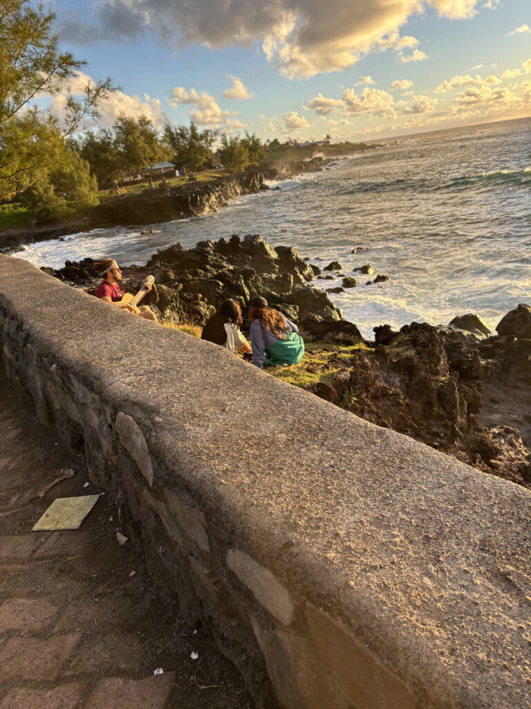 A scenic coastal view at sunset with a rocky shoreline and ocean waves. In the foreground, a stone wall runs along a path. Behind the wall, a person is sitting and playing a guitar, while two others are sitting nearby, enjoying the view. The sky is partly cloudy with a warm, golden light illuminating the scene. Trees and greenery are visible in the background along the coast.