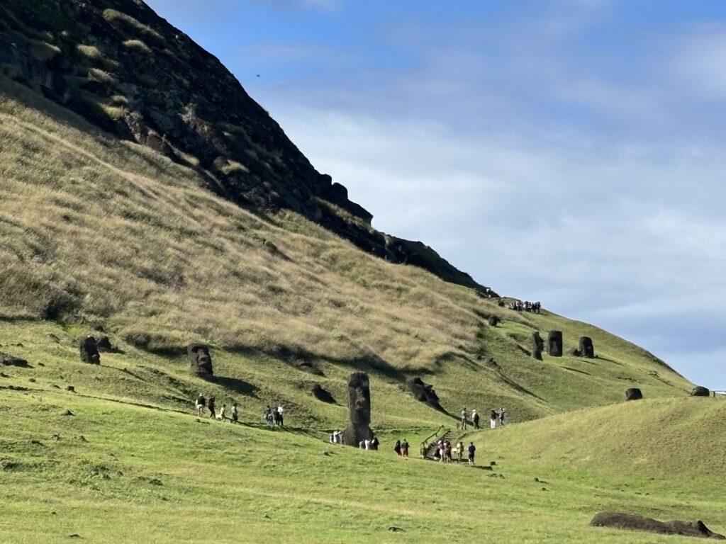 The image shows a grassy hillside with several large stone statues, known as Moai, on Easter Island. A group of people is walking along a path near the statues, and the sky is partly cloudy. The landscape is expansive, with the statues partially buried in the ground.