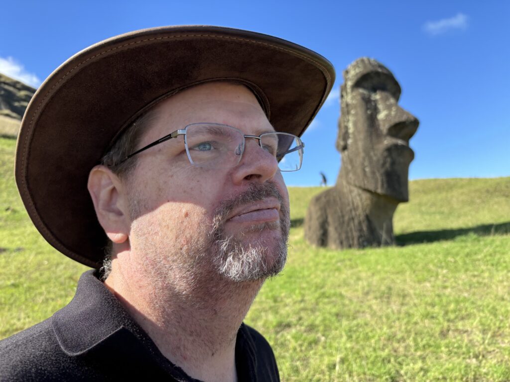 A man wearing a brown hat and glasses is looking into the distance. In the background, there is a large stone Moai statue on a grassy hill under a clear blue sky.