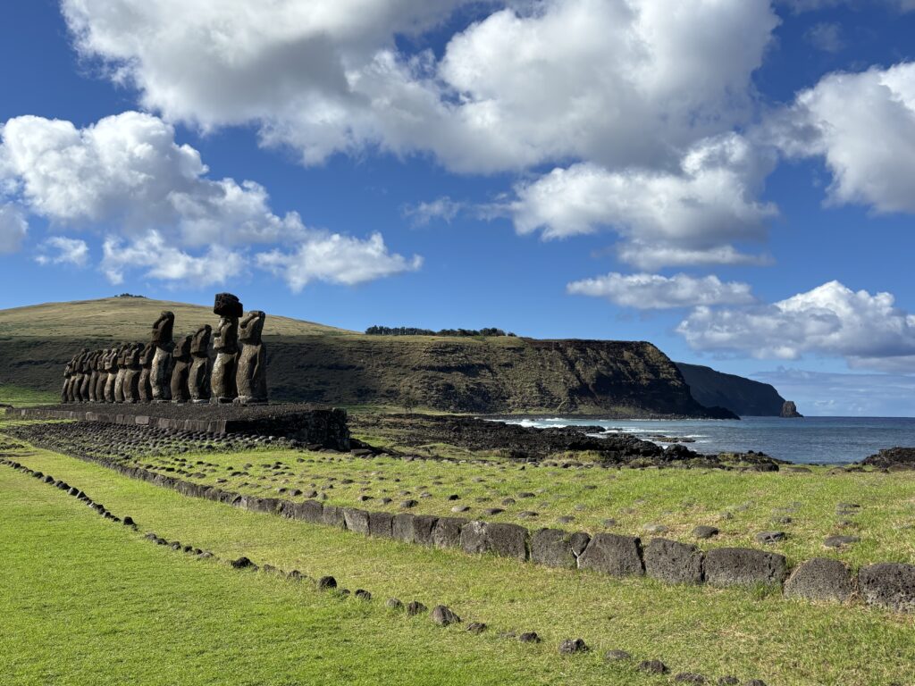 The image shows a row of large stone statues, known as moai, on a grassy landscape with a rocky coastline in the background. The sky is partly cloudy, and the ocean is visible to the right. The scene is likely from Easter Island.