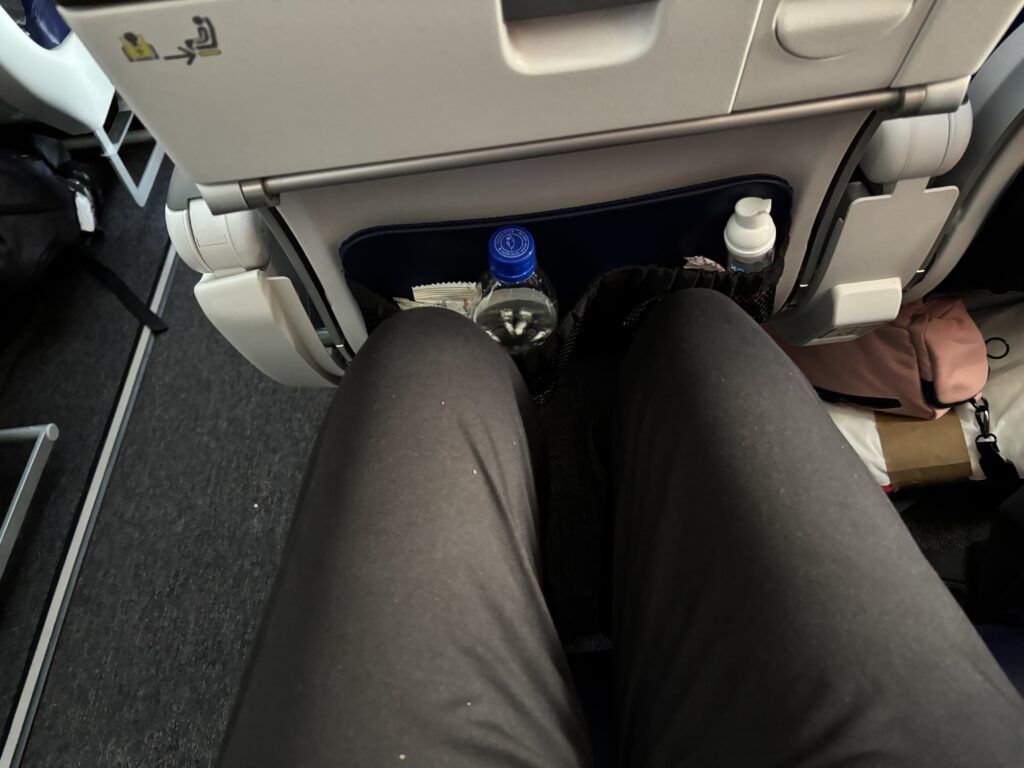 The image shows a view from an airplane seat, focusing on the legroom area. A person's legs are visible, wearing dark pants. The seatback pocket in front contains a water bottle and other small items. A pink bag is partially visible on the floor to the right. The airplane's interior, including the seat and tray table, is also visible.