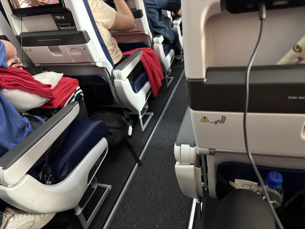 The image shows the interior of an airplane cabin, focusing on the seating area. There are several rows of seats with blue cushions and white armrests. Passengers are seated, and some have red blankets and pillows. The back of the seats in front have safety information and a screen. A bottle of water is visible in the seat pocket. The aisle is visible, and there is a charging cable plugged into the seat.