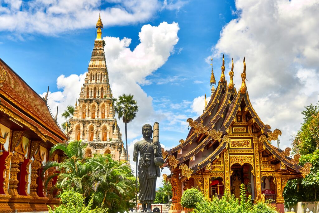 The image shows a vibrant scene of a traditional Thai temple complex. In the foreground, there is an ornate building with a richly decorated roof featuring intricate gold patterns and pointed spires. To the left, a tall pagoda with multiple tiers and niches containing statues is visible. A large statue of a standing figure is in the center, surrounded by lush green foliage. The sky is bright blue with scattered white clouds, adding to the serene atmosphere.