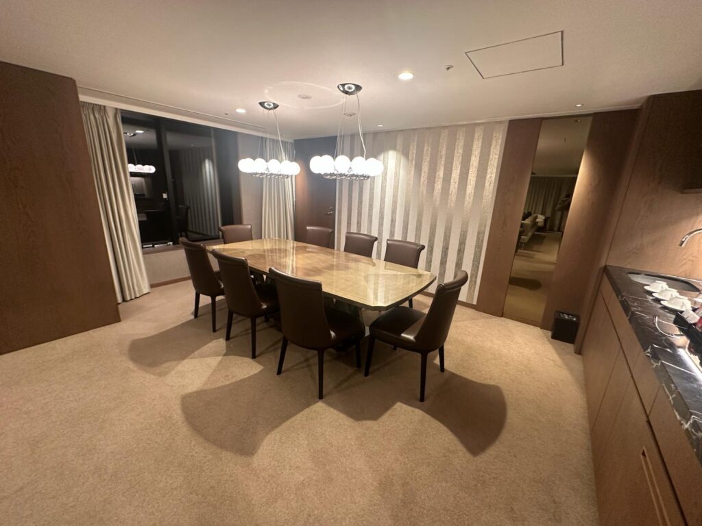 The image shows a modern dining room with a large rectangular table surrounded by eight brown chairs. Above the table, there are two contemporary chandeliers with spherical lights. The room has beige carpet flooring and is decorated with light-colored curtains and a striped accent wall. On the right side, there is a countertop with a sink and some dishes. The room appears spacious and well-lit.