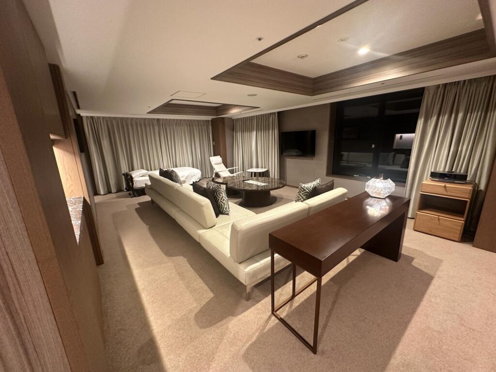 The image shows a modern, spacious living room with a beige carpet. There is a large white sectional sofa with patterned cushions facing a wall-mounted flat-screen TV. A round coffee table is in front of the sofa. Behind the sofa, there is a wooden console table with a decorative lamp. To the left, there is a bed with white linens, and a white chair is nearby. The room has large windows with closed curtains and a wooden cabinet with shelves on the right. The ceiling features recessed lighting.