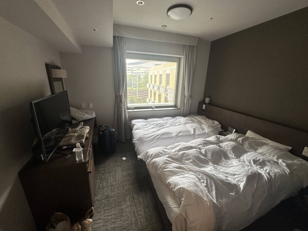 The image shows a hotel room with two unmade beds covered in white bedding. There is a window with curtains partially open, revealing a view of a building outside. A flat-screen TV is on a wooden dresser, which also holds some items like a water bottle and a bag. A suitcase is on the floor next to the dresser. The room has a neutral color scheme with gray walls and carpet.