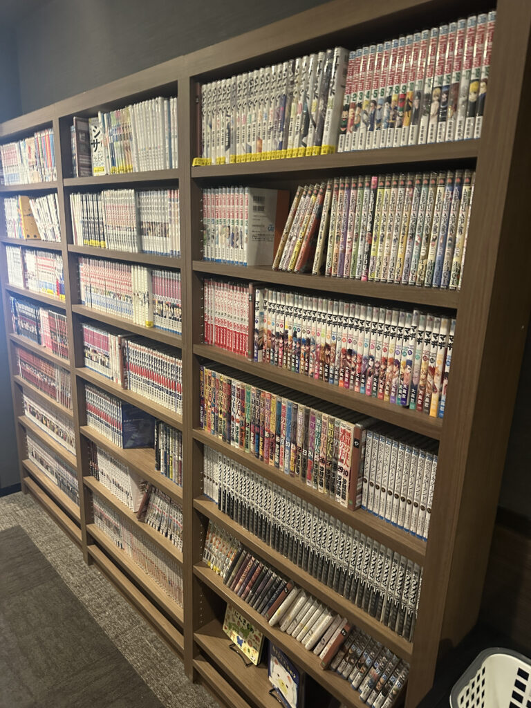 A large wooden bookshelf filled with neatly arranged manga volumes. The shelves are packed with various series, each with colorful spines displaying different titles and artwork. The room has a carpeted floor, and there is a laundry basket visible at the bottom right corner.