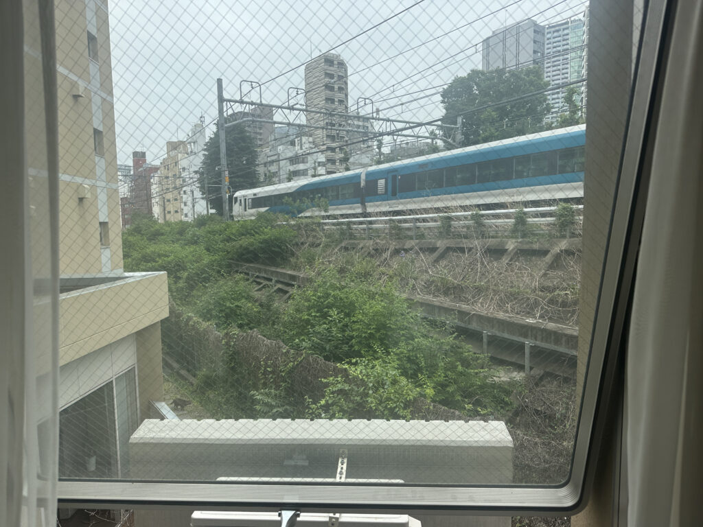The image shows a view through a window with a mesh screen. Outside, there is a train with a blue and white color scheme traveling on elevated tracks. The surrounding area includes greenery and several tall buildings in the background.