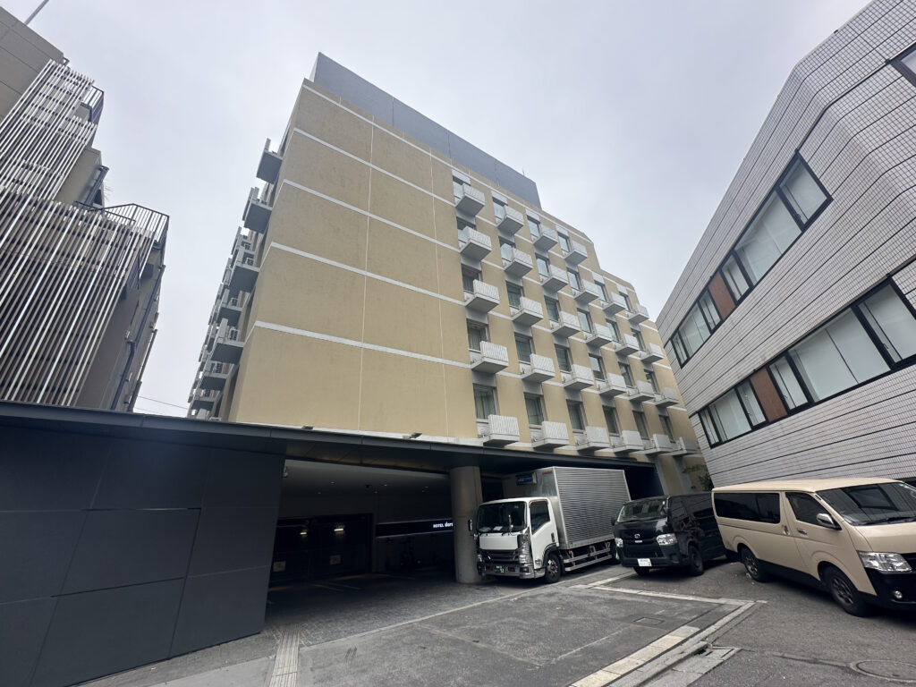 The image shows a street view of a modern urban area with several buildings. The central building is a multi-story structure with a beige facade and small balconies. To the right, there is a white building with horizontal lines and large windows. In front of the buildings, there are three parked vehicles: a white truck, a black van, and a beige van. The sky is overcast, giving the scene a muted appearance.
