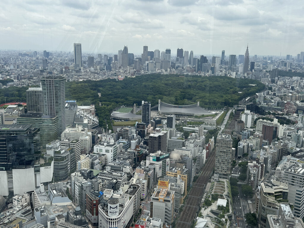 Aerial view of a large cityscape featuring numerous high-rise buildings and dense urban development. In the center, there is a large green park area with a distinctive, curved-roof building. The skyline is filled with various skyscrapers, and the sky is overcast with clouds.