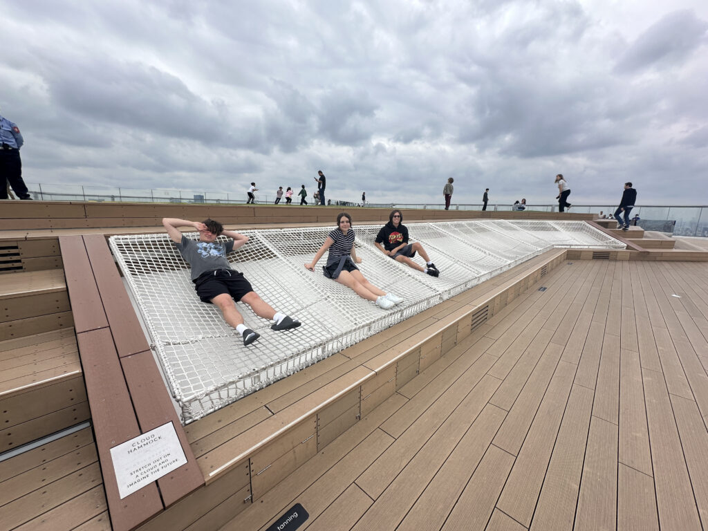 The image shows three people relaxing on a large, white net hammock structure on a rooftop deck. The deck is made of light brown wood, and there are several other people walking or standing in the background. The sky is overcast with gray clouds. A sign next to the hammock reads "Cloud Hammock."