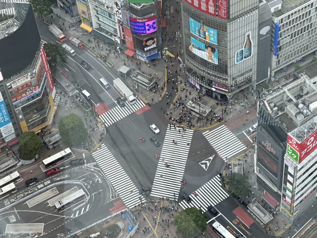 Aerial view of a busy urban intersection, likely Shibuya Crossing in Tokyo, Japan. The image shows multiple crosswalks converging, with numerous pedestrians crossing in various directions. Surrounding the intersection are tall buildings with colorful advertisements and billboards. Several vehicles, including buses and cars, are visible on the roads. The scene is bustling with activity, typical of a vibrant city center.