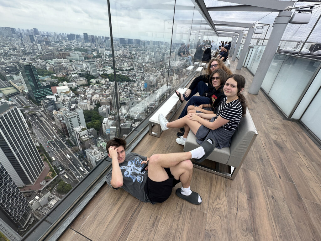 A group of people is sitting and lying on a rooftop observation deck with a glass barrier, overlooking a sprawling cityscape. The deck has wooden flooring, and the city below is filled with tall buildings and roads. The sky is overcast, and the people appear relaxed, enjoying the view.