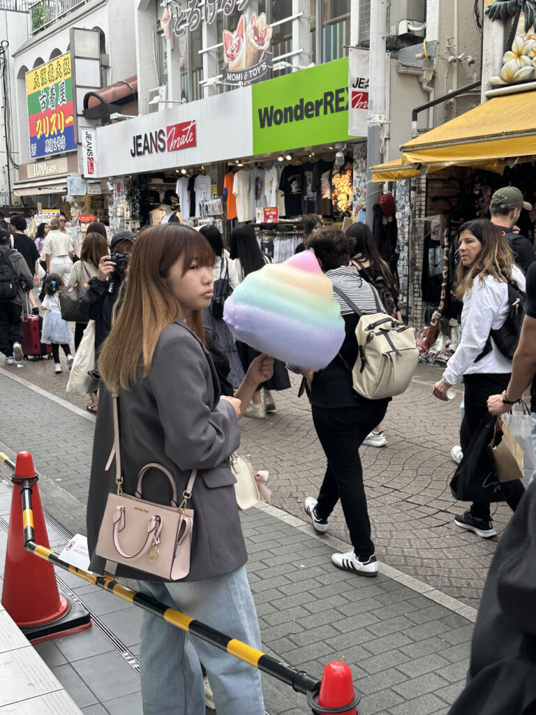 A woman is standing on a busy street holding a large, colorful cotton candy. She is wearing a gray jacket and carrying a light pink handbag. The street is crowded with people walking past various shops, including one with a sign that reads "JEANS Mate" and another with "WonderREX." The atmosphere is lively, with many pedestrians and storefronts visible.