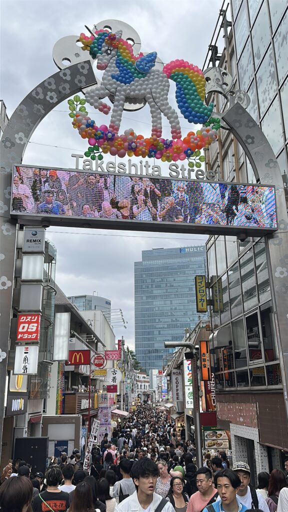 The image shows the entrance to Takeshita Street in Harajuku, Tokyo. A large decorative archway features a colorful unicorn made of balloons. Below the arch, a digital screen displays a crowd of people. The street is bustling with many pedestrians, and various shop signs are visible on both sides. In the background, modern buildings rise against a cloudy sky.