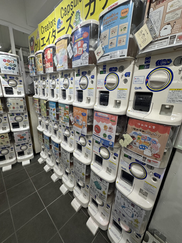 The image shows a row of capsule toy vending machines, also known as gashapon, in a store. Each machine has a display showing various toy options available inside the capsules. The machines are white with coin slots and turn knobs. There are colorful posters and signs above the machines, featuring illustrations and text in Japanese.