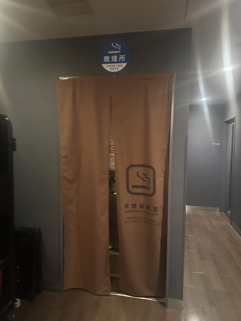 The image shows an entrance to a designated smoking room. There is a brown curtain with a smoking symbol and text in multiple languages, including English and Japanese, indicating it is a smoking area. Above the entrance, there is a blue sign with a smoking symbol and the words "Smoking Area" in English and Japanese. The surrounding area has a modern interior with a wooden floor.