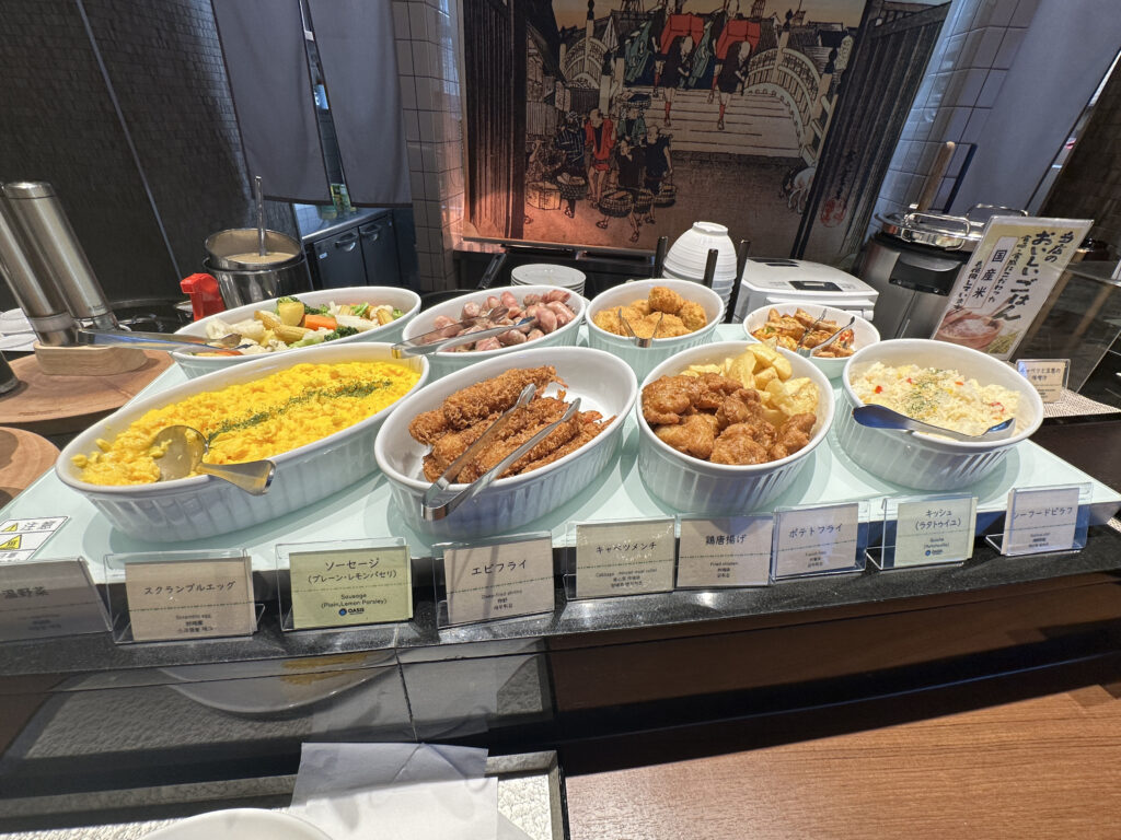 The image shows a buffet setup with various dishes in white serving dishes. The dishes include scrambled eggs, sausages, fried items, rice, and vegetables. Each dish has a label in front of it, providing names and descriptions. The background features a decorative wall with an illustration, and there are utensils and serving tools placed around the dishes.