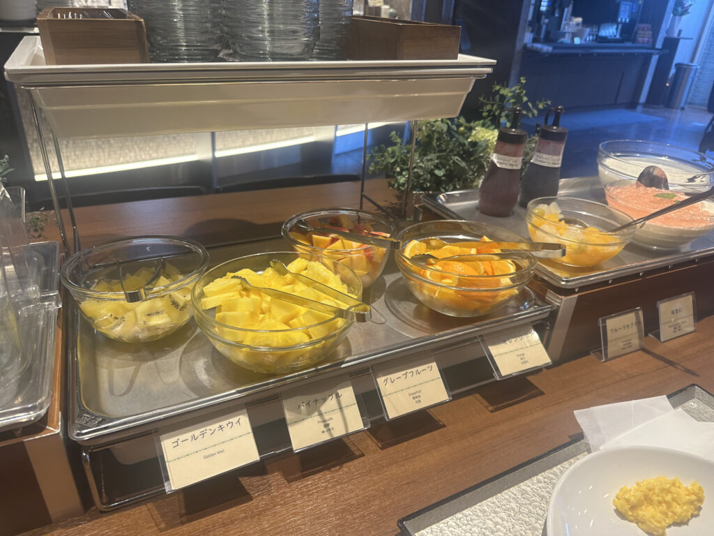 The image shows a buffet setup with various bowls of sliced fruits. From left to right, there are bowls containing golden kiwi, pineapple, grapefruit, and a fruit cocktail. Each bowl has a serving tong. Behind the fruit, there are bottles of sauces or dressings. In the foreground, there is a plate with a small serving of scrambled eggs. The setting appears to be a breakfast buffet in a restaurant or hotel.
