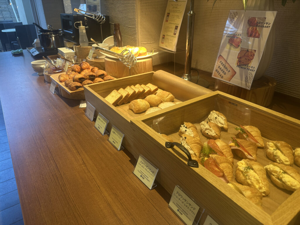 The image shows a buffet-style setup with a variety of baked goods displayed on a wooden counter. There are trays filled with different types of bread, croissants, and pastries. Some items are labeled with small signs in front of them. The setup includes a soup pot, a toaster, and condiment bottles. There are also posters on the wall advertising freshly baked bread and croissants. The setting appears to be a breakfast or brunch buffet in a restaurant or hotel.