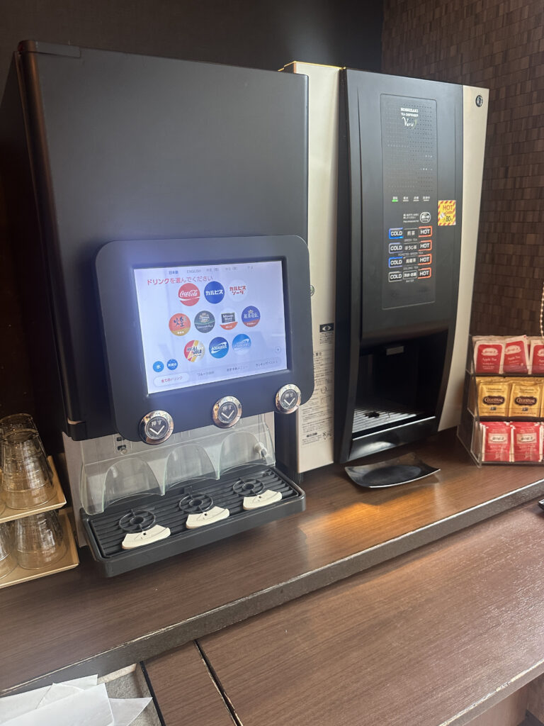 The image shows a beverage station with two drink dispensers. The dispenser on the left has a touchscreen interface displaying various drink options, including Coca-Cola and other beverages, with buttons below for selection. The dispenser on the right has buttons labeled for hot and cold drinks. To the side, there are stacked plastic cups and a container with packets, likely for tea or coffee. The setup is on a wooden counter.