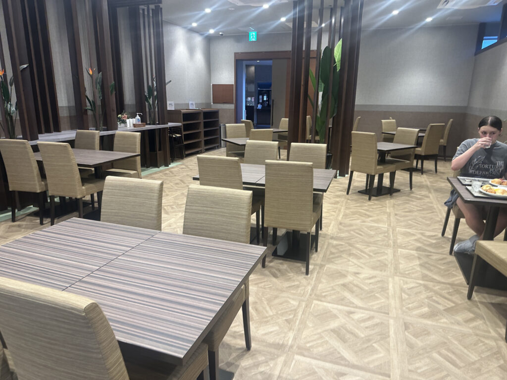 The image shows a dining area with several empty tables and chairs arranged neatly. The room has a modern design with wooden accents and decorative plants. There is one person sitting at a table on the right side, eating and drinking. The lighting is bright, and the floor has a patterned design.