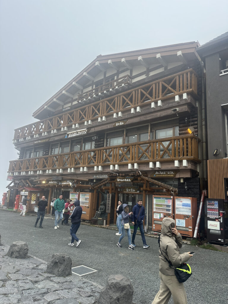 A wooden building with multiple balconies and a sloped roof, featuring signs in Japanese. Several people are walking in front of the building, some wearing jackets and hoodies, suggesting cool weather. There are vending machines and a statue near the entrance. The sky is overcast and foggy, creating a misty atmosphere.