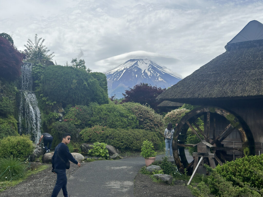 A scenic landscape featuring a snow-capped mountain in the background, partially covered by clouds. In the foreground, there is a lush garden with a small waterfall cascading over rocks. A traditional wooden waterwheel is on the right side, attached to a thatched-roof structure. Two people are visible: one walking on a path and another near the waterfall. The scene is serene and natural.
