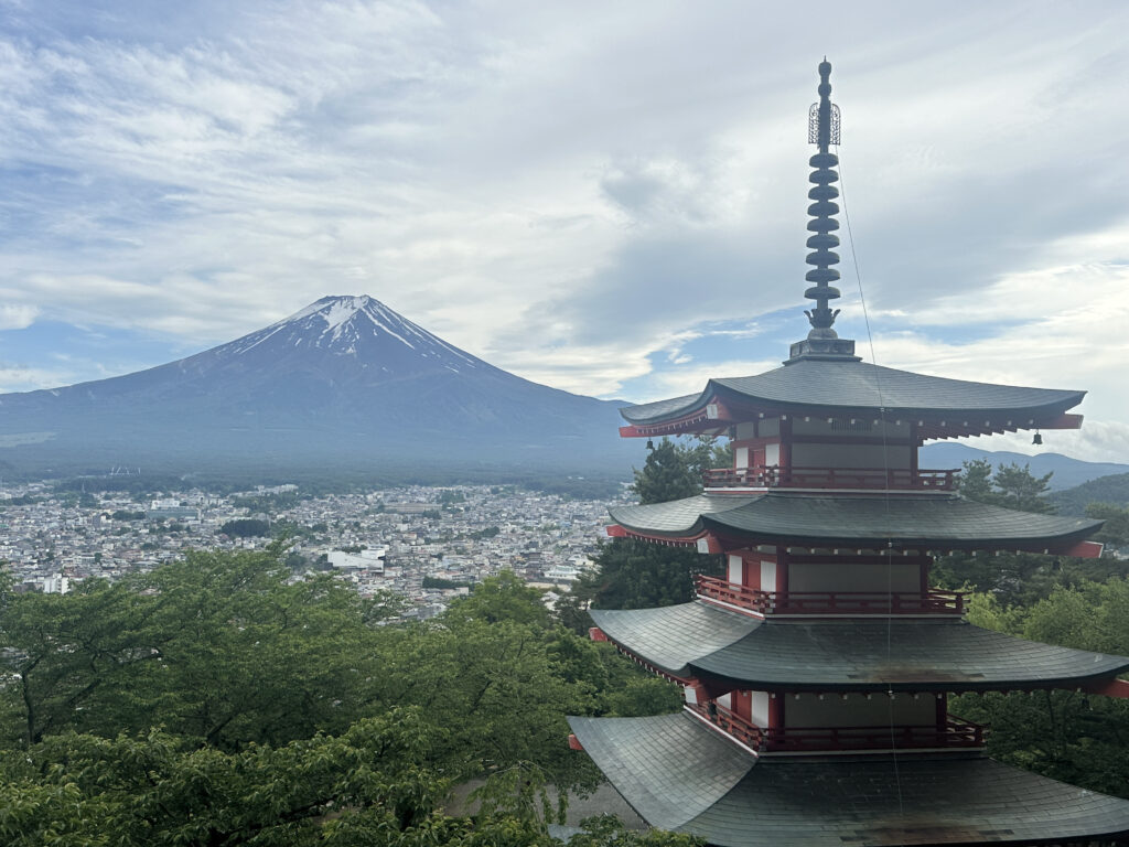 A scenic view featuring a traditional Japanese pagoda in the foreground, with a cityscape and lush green trees below. In the background, Mount Fuji rises majestically under a partly cloudy sky.