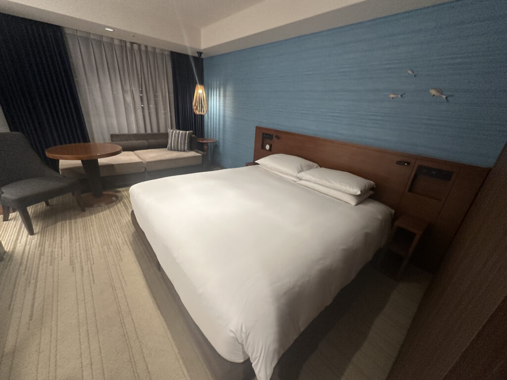 The image shows a modern hotel room with a large bed covered in white linens. The headboard is wooden, and the wall behind it is painted blue with small decorative elements. To the left, there is a seating area with a gray chair, a round wooden table, and a beige sofa with a striped pillow. A unique hanging light fixture is above the table. The room has a carpeted floor and curtains covering the window.