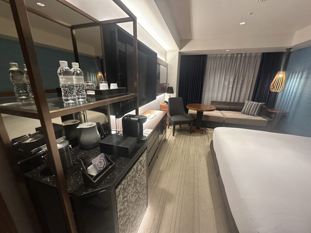 The image shows a modern hotel room with a neatly made bed on the right. To the left, there is a shelf with bottled water, a coffee maker, and other amenities. In the background, there is a seating area with a sofa, a round table, and a chair. The room has a large window with curtains and a stylish hanging light fixture. The decor is contemporary with neutral tones.