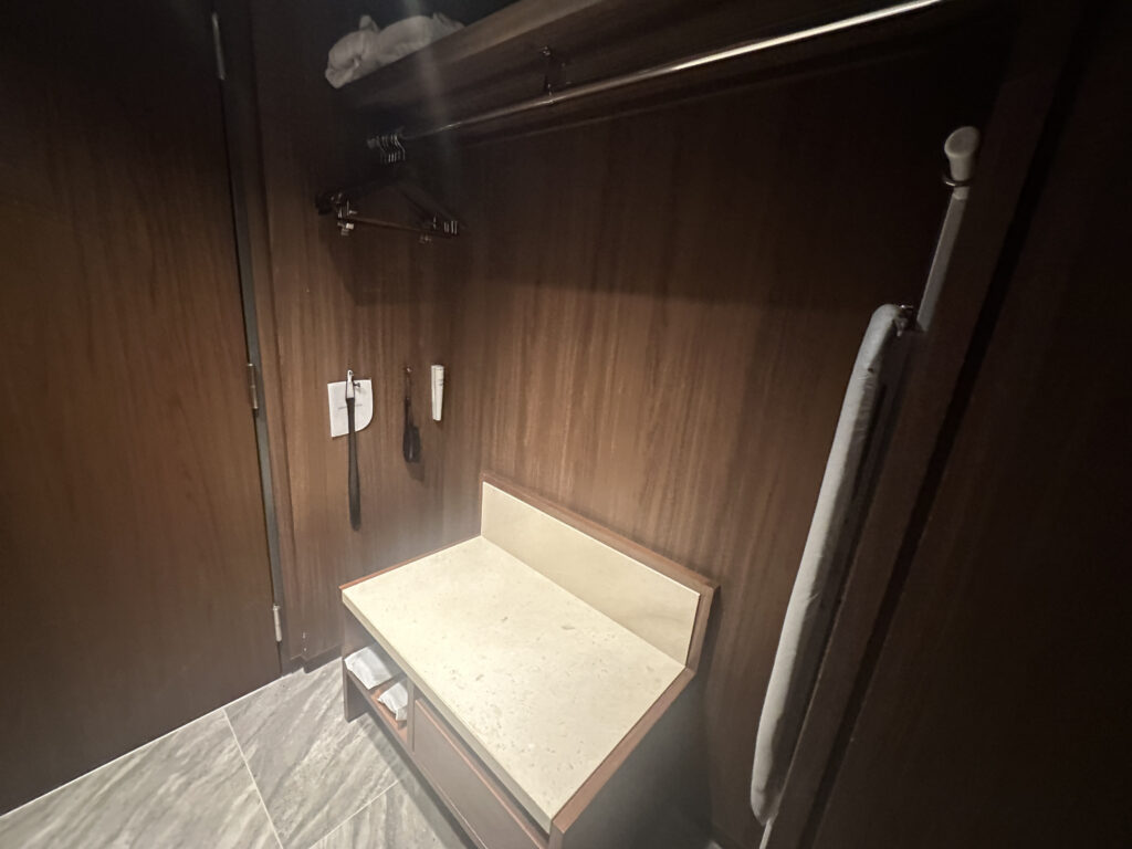 The image shows a wooden closet interior with a shelf and a hanging rod. There are a few hangers on the rod, and a folded towel is placed on the shelf. Below, there is a bench with a light-colored surface. An ironing board is visible on the right side, and a couple of items, possibly a shoehorn and a clothes brush, are hanging on the wall. The floor is tiled.