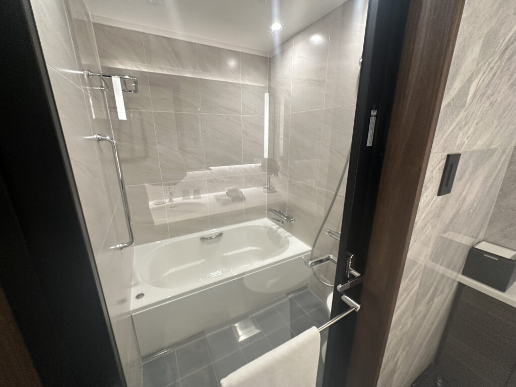 The image shows a modern bathroom with a bathtub. The walls are tiled with light-colored tiles, and there is a glass partition separating the bathtub from the rest of the bathroom. A chrome showerhead and faucet are visible, along with a towel hanging on a rail attached to the glass door. The floor is covered with dark tiles.