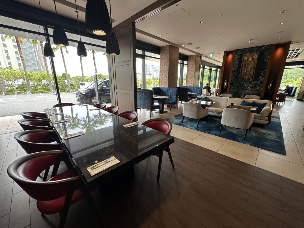 The image shows a modern, stylish restaurant interior with a mix of seating options. In the foreground, there is a long, dark marble table with red chairs. In the background, there are several seating areas with sofas and armchairs on a blue rug. Large windows line the walls, allowing natural light to illuminate the space. Outside, palm trees and a parked car are visible. The decor is contemporary, with pendant lights hanging above the table.