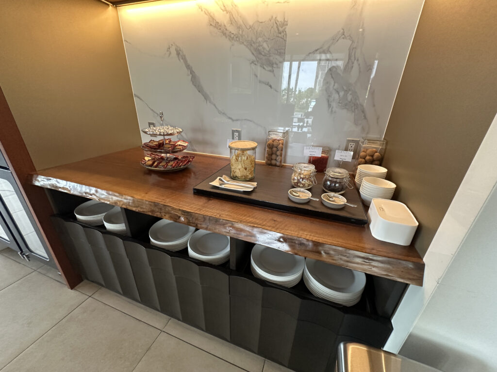 The image shows a wooden countertop with a variety of snacks and condiments. On the left, there is a tiered stand with packaged snacks. In the center, there are jars containing different items, including cookies and possibly nuts or dried fruits. There are also small bowls with spoons on a tray. On the right, there are stacks of small bowls and a white container. Below the counter, there are shelves with stacks of plates. The background features a marble-patterned wall.