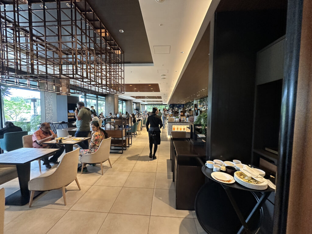 The image shows a modern restaurant interior with several people dining. There are tables and chairs arranged throughout the space, with some patrons seated and eating. A server is walking through the restaurant. The decor includes a geometric ceiling design and large windows that let in natural light. A stack of used plates and cups is visible on a cart to the right.