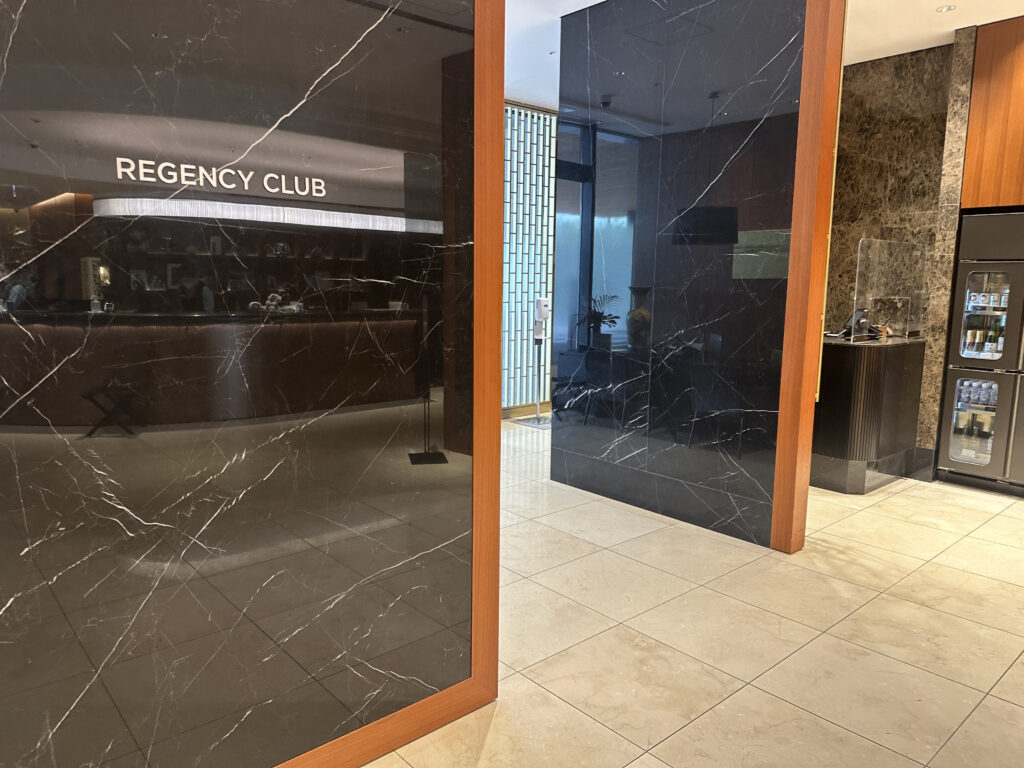 The image shows the entrance to a "Regency Club" with a modern and elegant interior. The walls are made of dark marble with white veining, and there is a reflective surface displaying the club's name. The floor is tiled in a light color. To the right, there is a small counter with a transparent protective screen and a refrigerator stocked with beverages. The overall atmosphere is upscale and sophisticated.