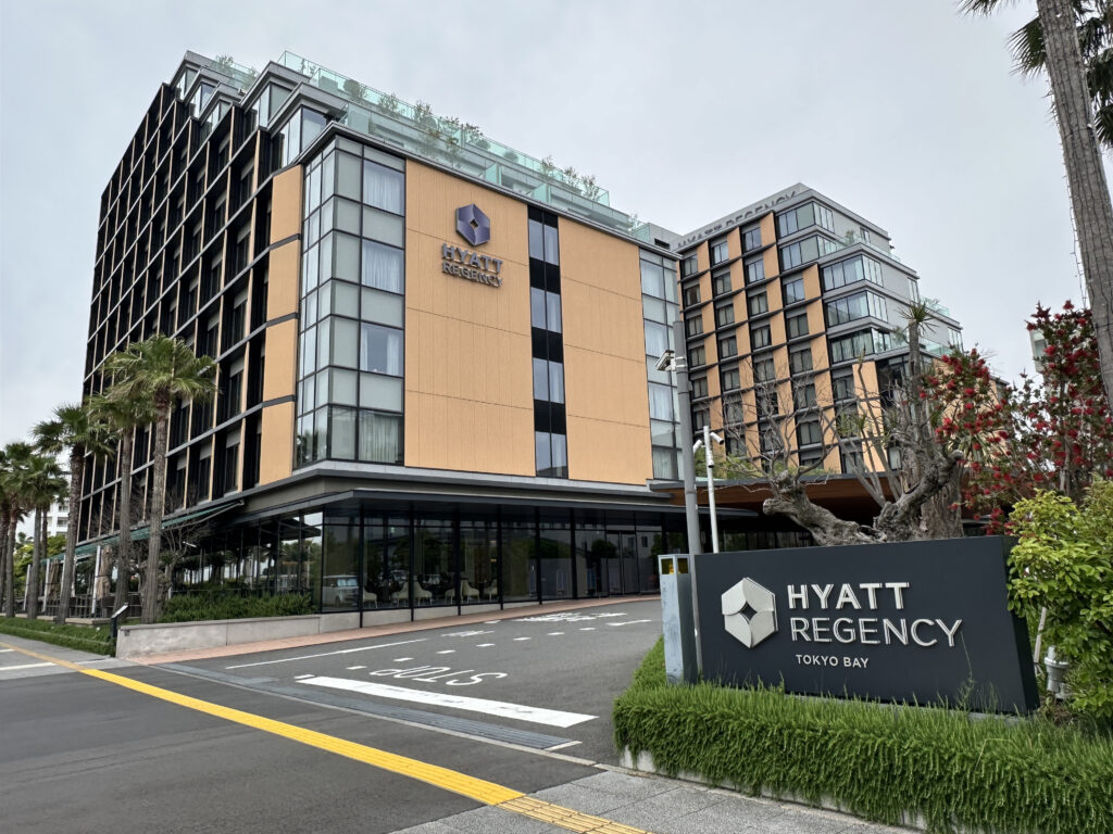 The image shows the exterior of the Hyatt Regency Tokyo Bay hotel. The building is modern, with large glass windows and a combination of beige and black paneling. In the foreground, there is a sign with the hotel’s name and logo, surrounded by greenery and trees. The sky is overcast.