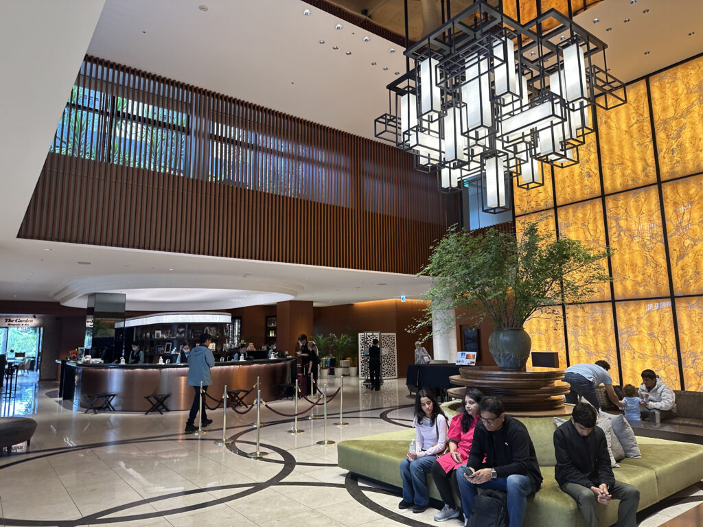 The image shows a hotel lobby with a modern and elegant design. There is a large, geometric chandelier hanging from the ceiling. A group of people is seated on a circular green sofa with a large potted plant in the center. In the background, there is a bar area with people standing and sitting. The walls have a warm, textured design, and the floor is tiled with a circular pattern.