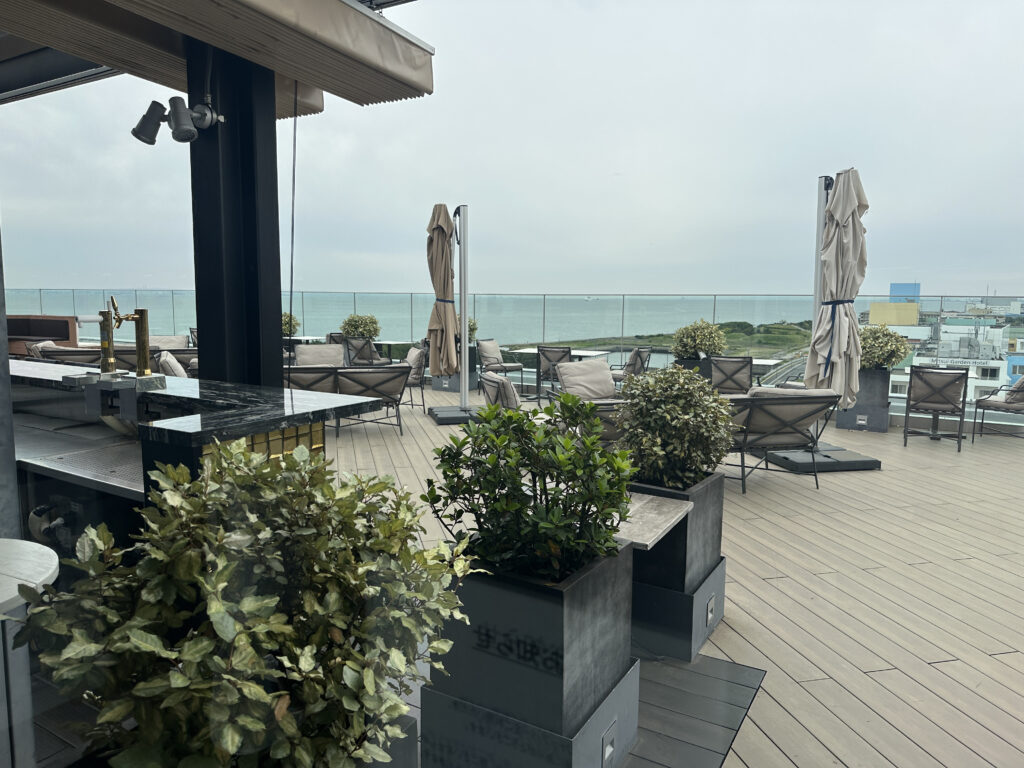 A rooftop terrace overlooking the ocean, featuring a bar area with a countertop and beer taps. The terrace has several lounge chairs and tables, with potted plants and closed umbrellas scattered around. The sky is overcast, and the ocean is visible in the background.