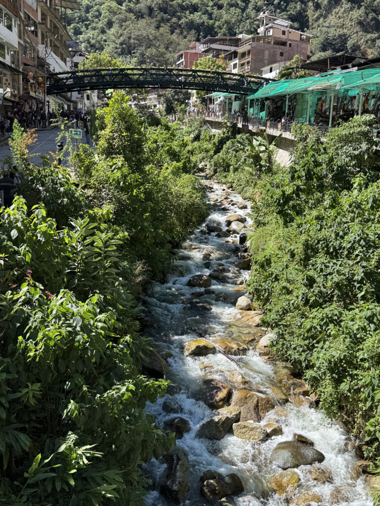 A scenic view of a small, rocky river flowing through a lush, green area in a town. On the left, there are buildings and a street with people walking. A green metal bridge crosses over the river, connecting both sides. The background features more buildings and a forested hillside. The scene is vibrant and sunny.